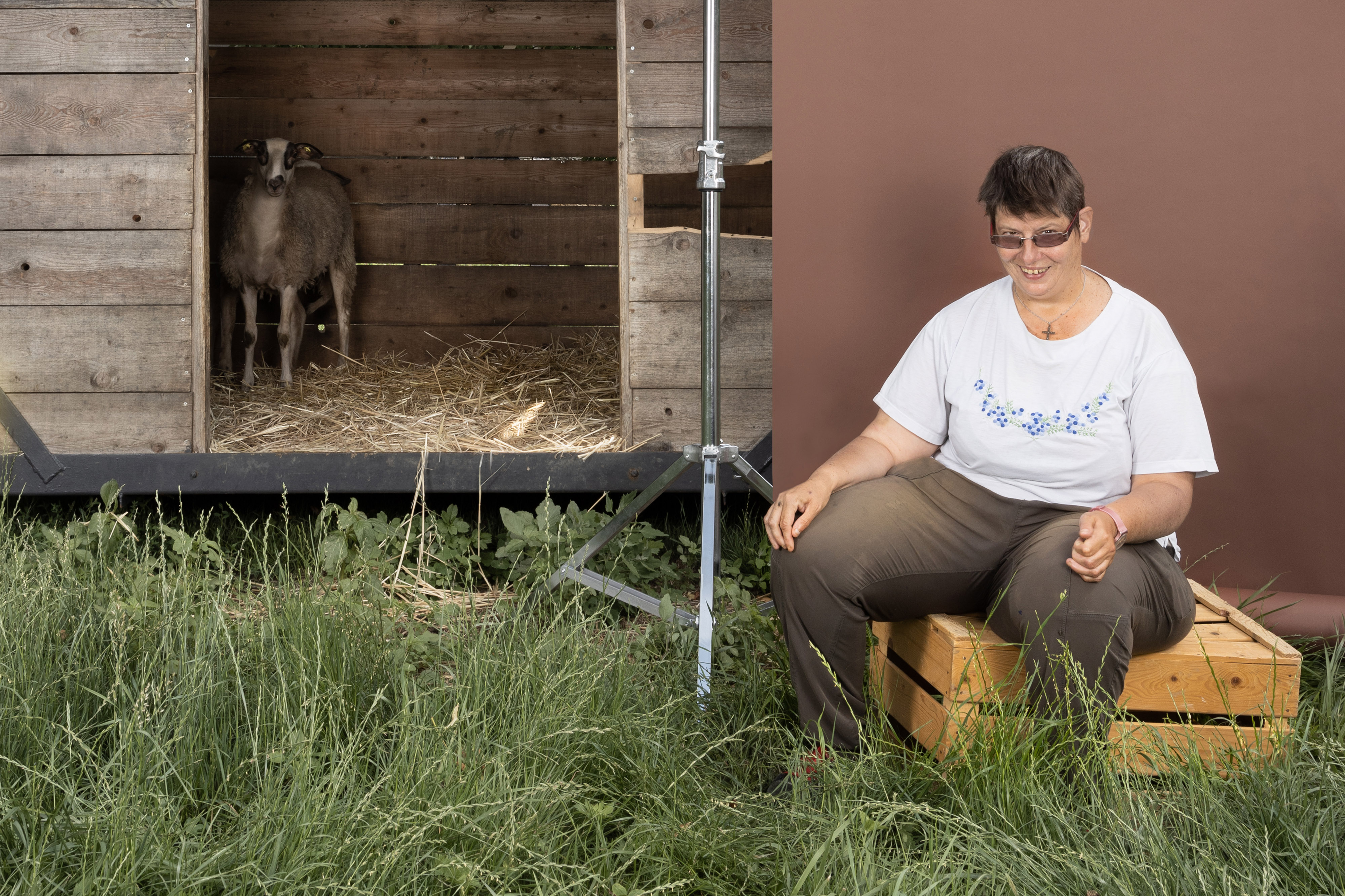 Ein Foto aus dem Buch "Ohne Milch und Zucker". Rechts sitzt Nicole Leuenberger auf einer Holzkiste vor einer brauenen Leinwand, links daneben sieht man ein Schaf.