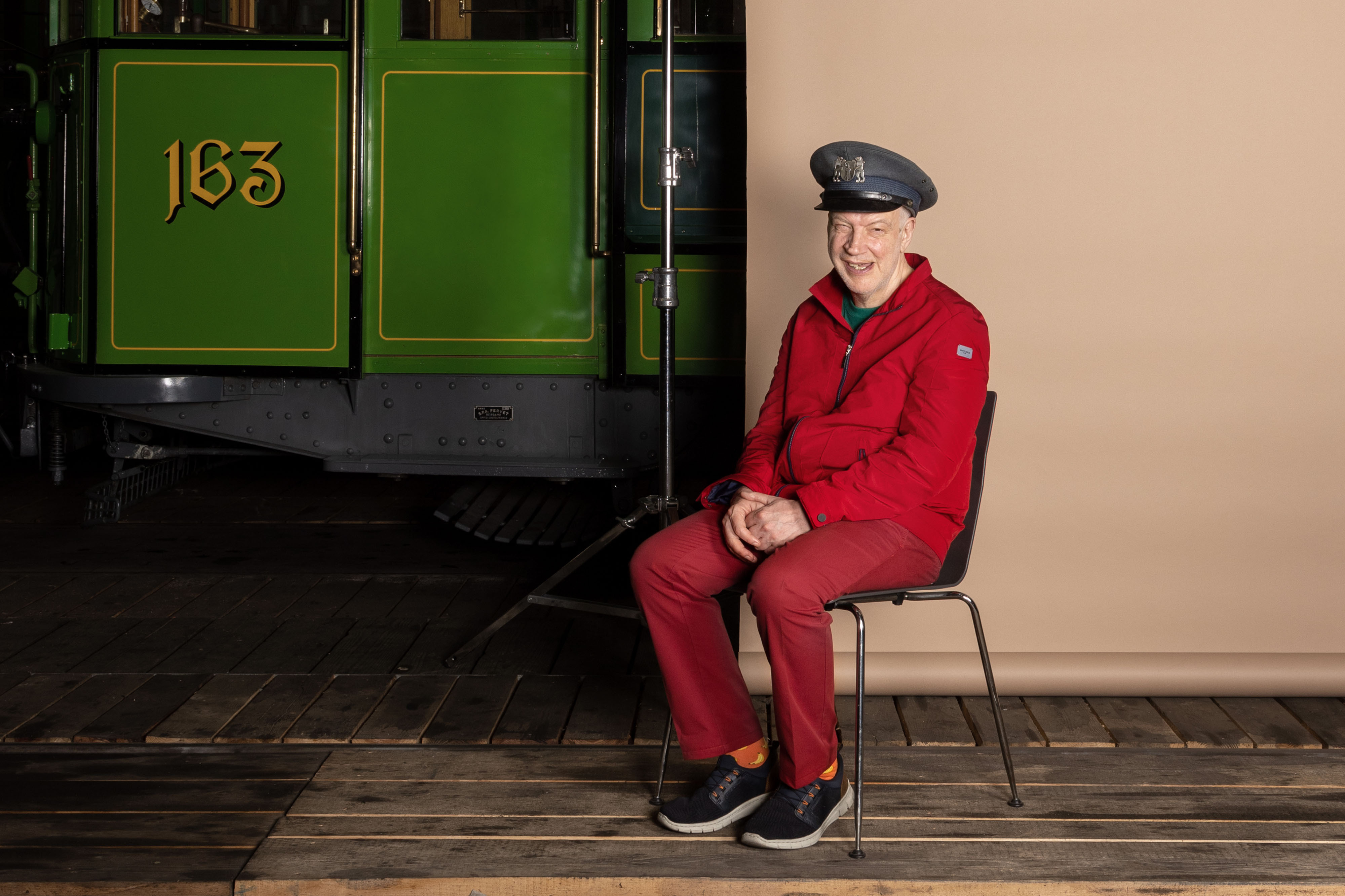 Ein Foto aus dem Buch "Ohne Milch und Zucker". Auf der rechten Seite sitzt Pius auf einem Stuhl vor einer beigen Leinwand, links sieht man einen Teil eines alten Trams.